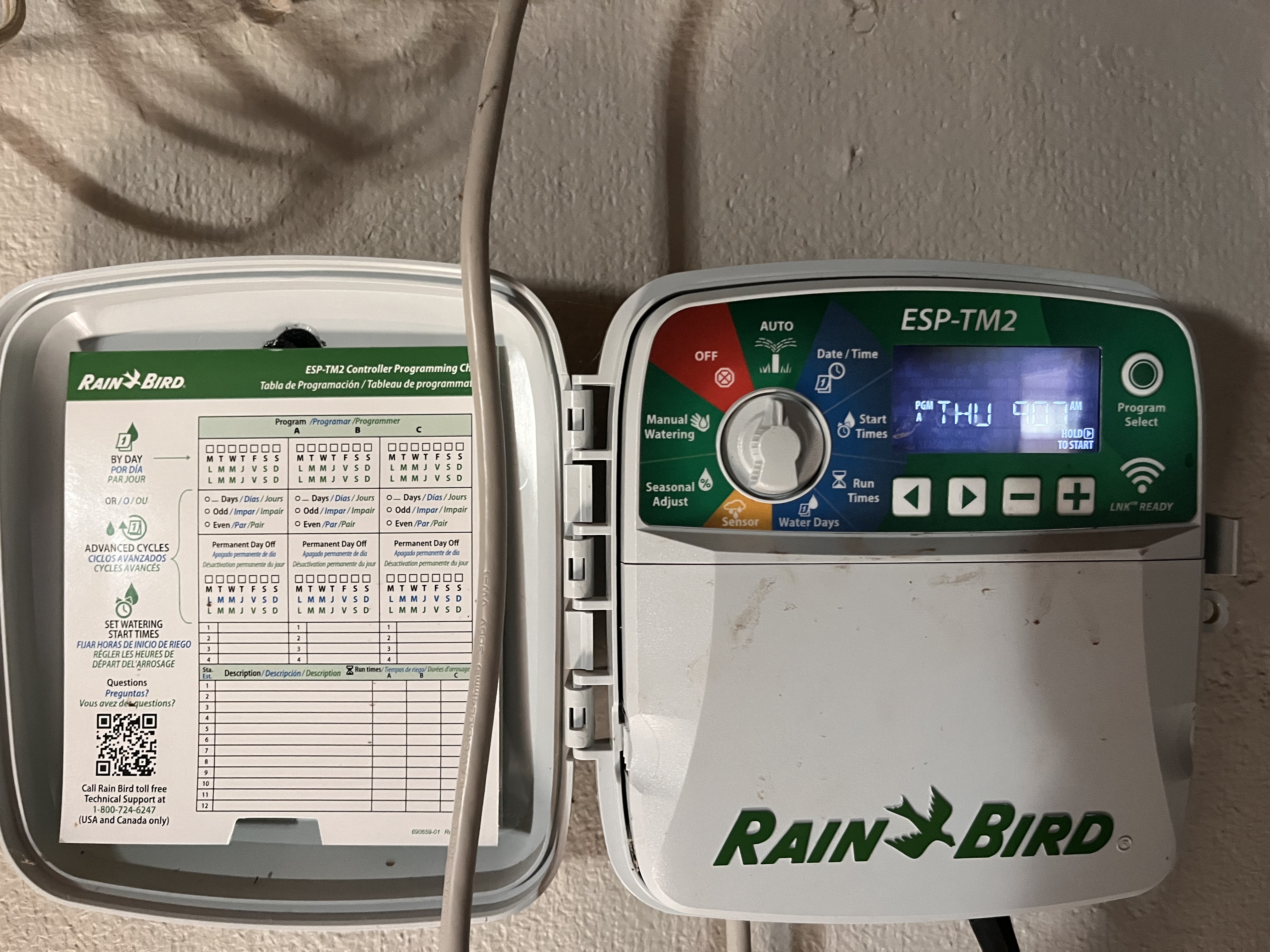 A Rain Bird sprinkler controller with a dial and buttons, next to a paper programming chart taped inside the door