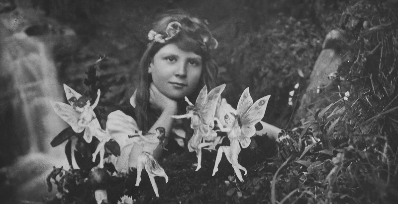 Black-and-white Cottingley Fairies photograph of a young girl with flowers in her hair gazing calmly at the camera while four paper cut-out fairies appear to dance in the grass in front of her